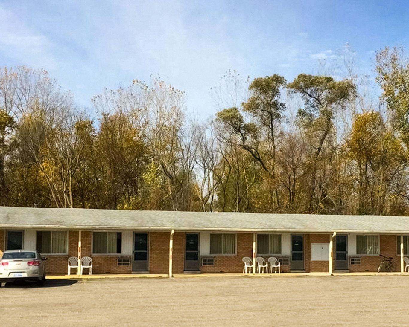Single-story brick motel with outdoor seating and autumn trees in the background.