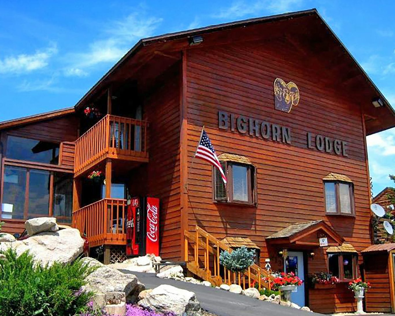 Rustic exterior of Bighorn Lodge with American flag and Coca-Cola vending machine under clear blue sky.