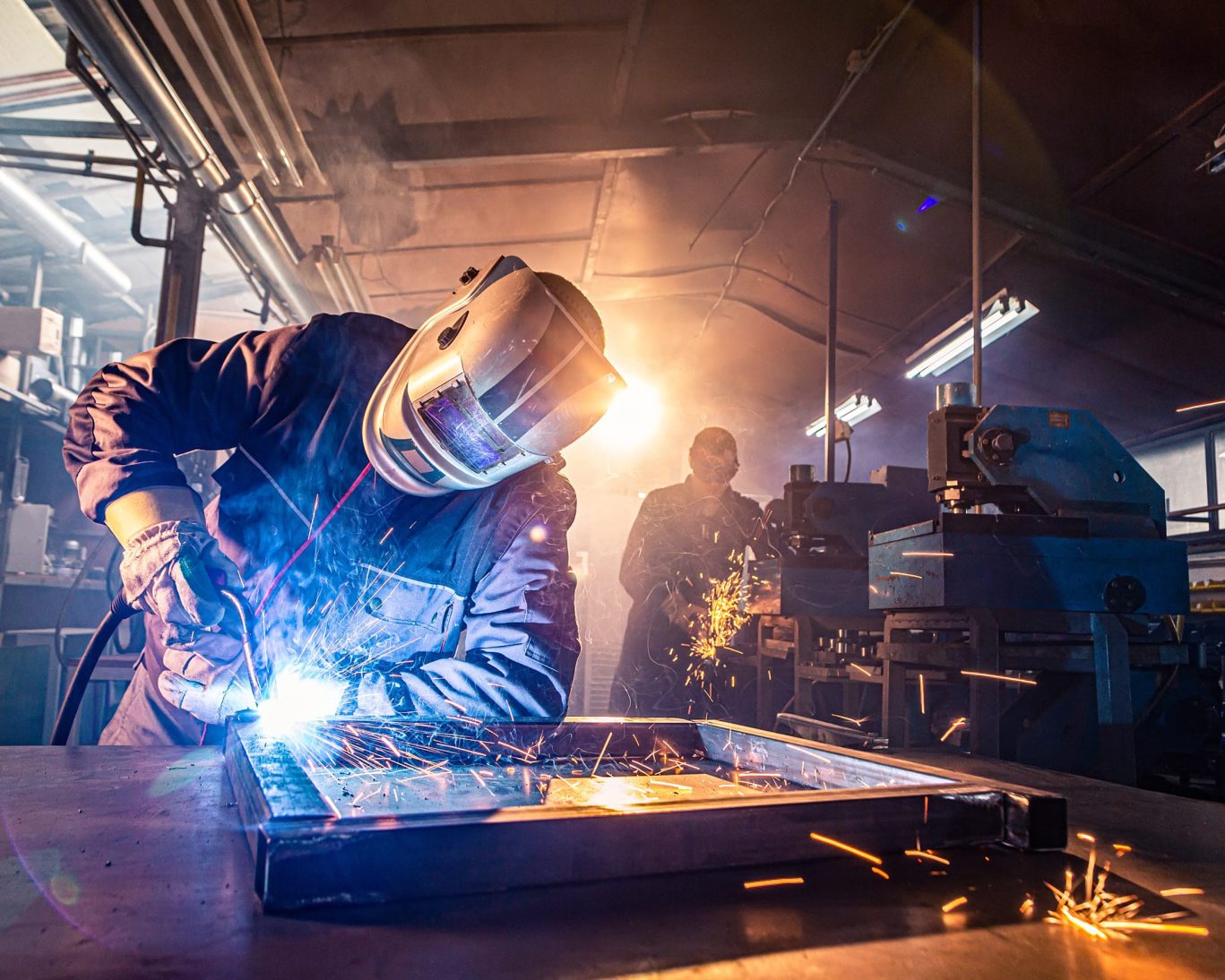 Worker welding metal frame at Warren Manufacturing facility with bright sparks and protective gear.