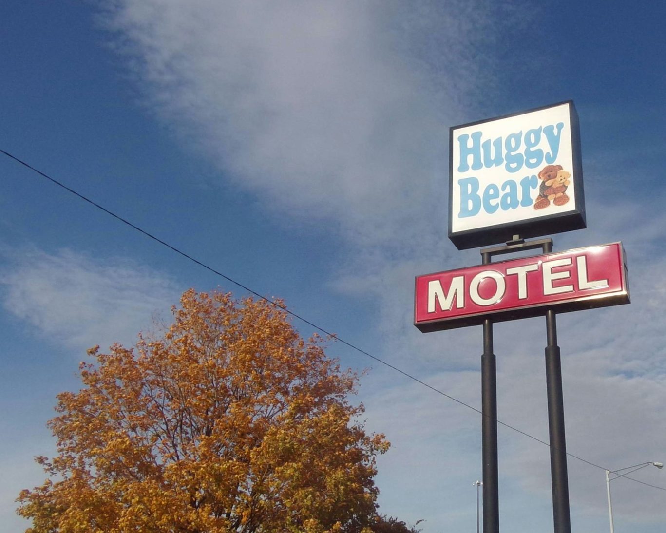 Huggy Bear Motel roadside sign with teddy bear logo under blue sky and fall foliage.