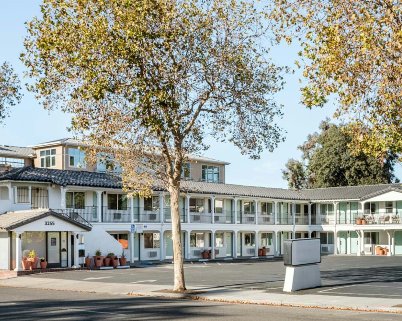 Front exterior of a two-story white motel building with light blue doors and tree-lined street.