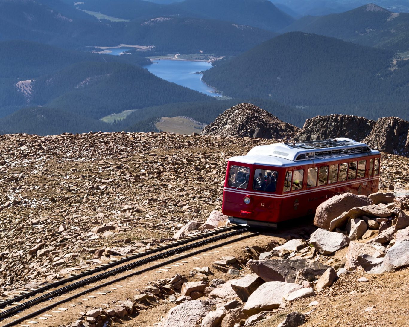 Red cog railway train ascending rocky terrain of Pikes Peak with panoramic views of lakes and mountains.