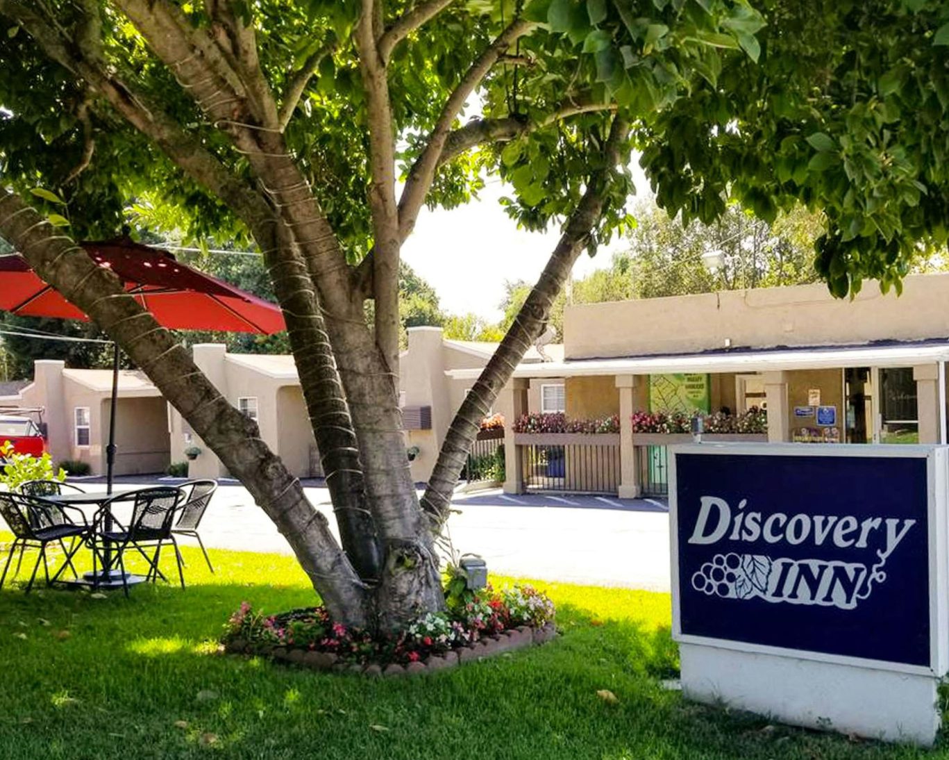 Front view of Discovery Inn Napa Valley with tree-shaded seating area and hotel signage in a garden setting.