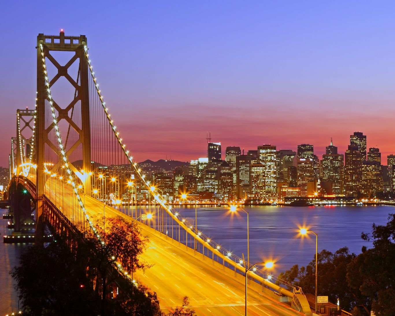 Bay Bridge illuminated at twilight with the San Francisco skyline glowing in the background.