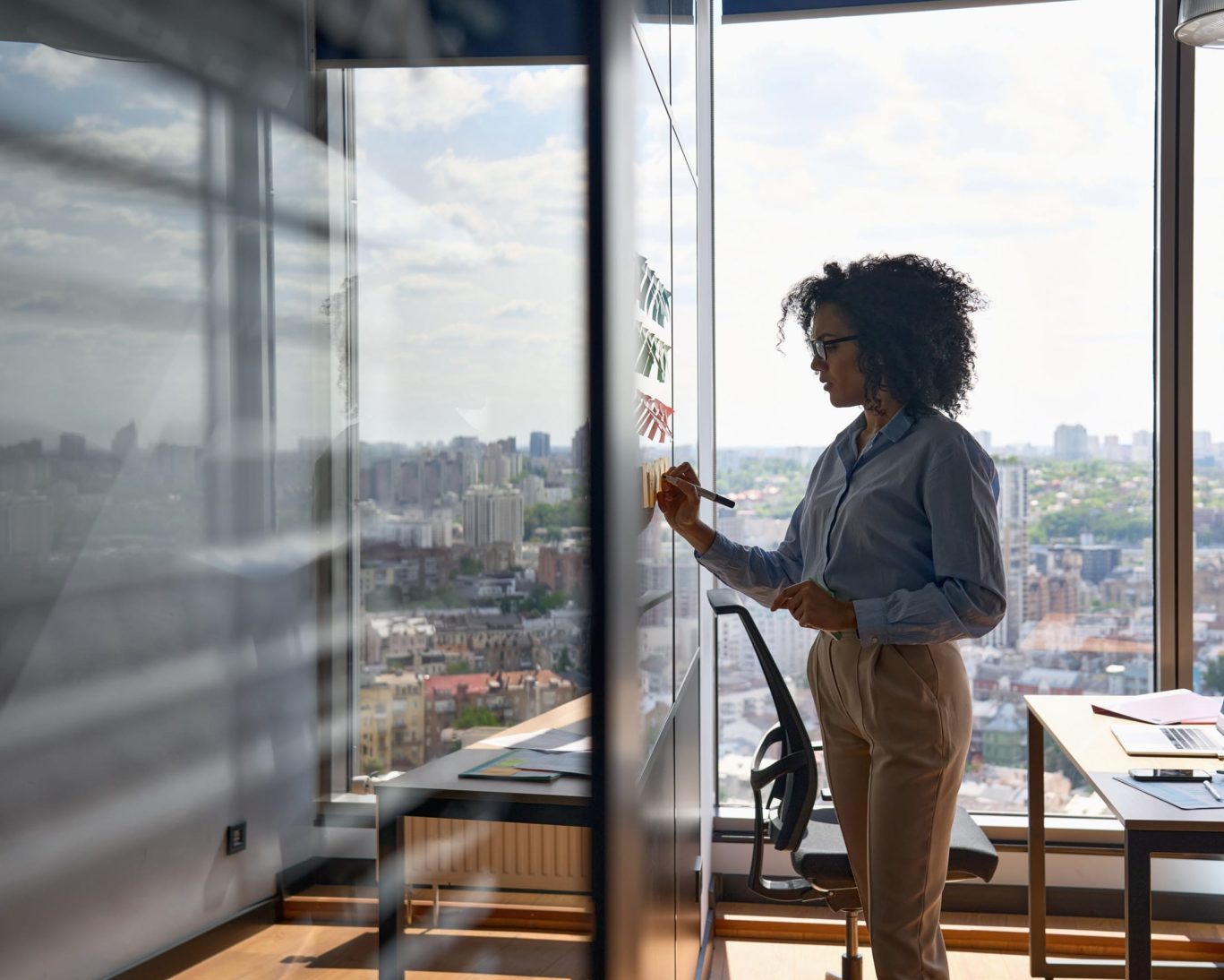 Professional businesswoman writing on a glass wall with panoramic city views from a modern office.