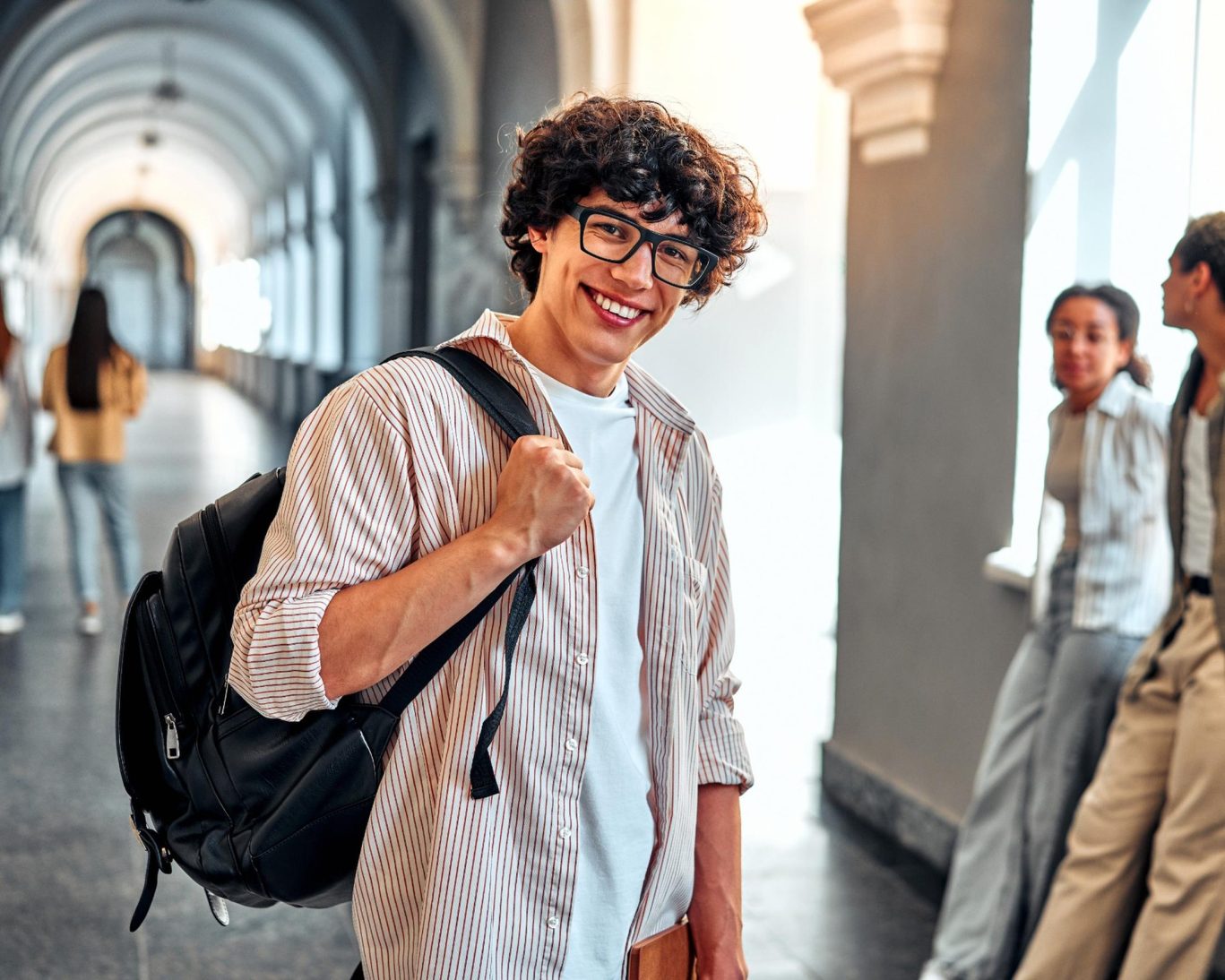 Smiling university student walking through a campus hallway with a backpack and books.