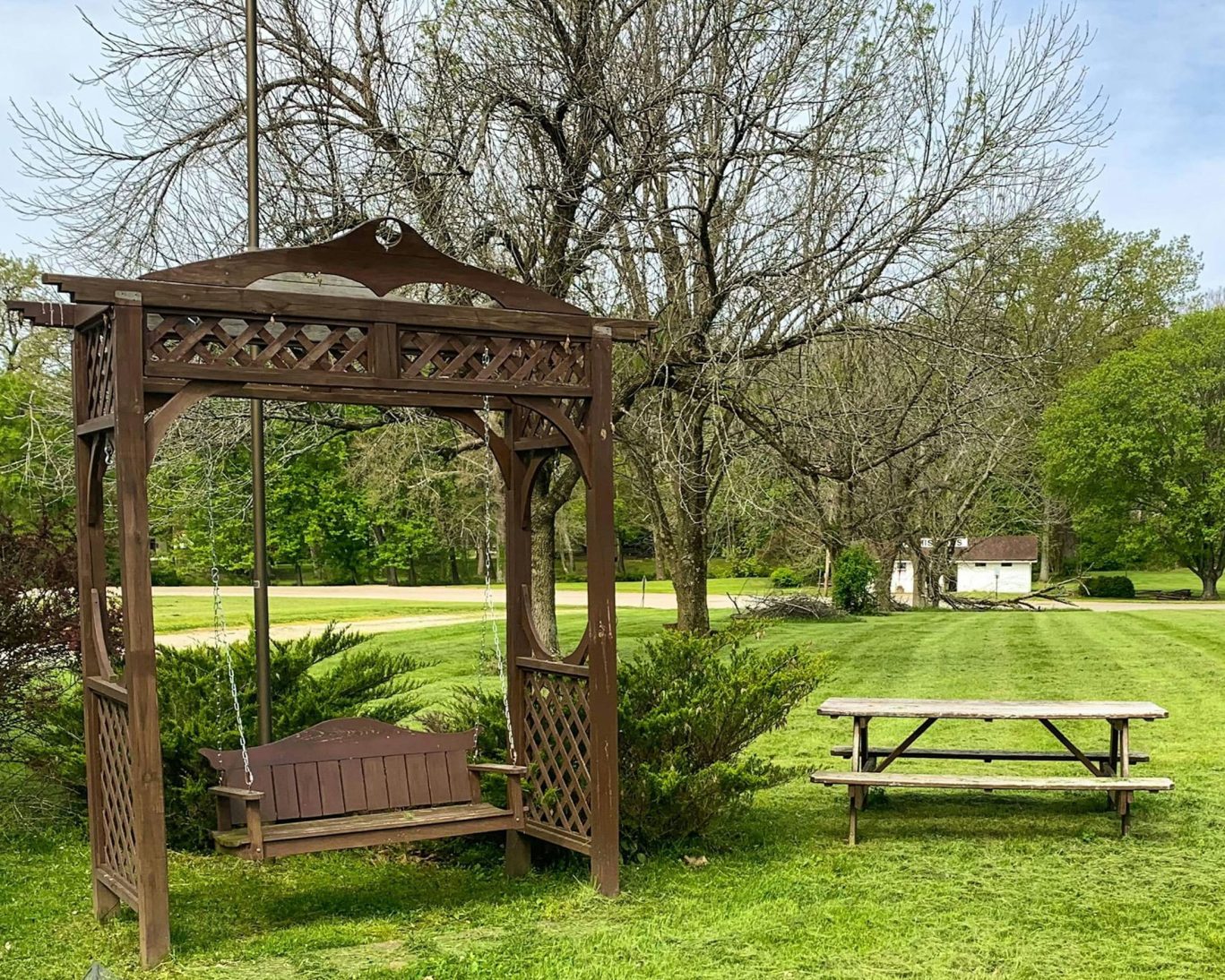 Wooden porch swing and picnic bench in grassy outdoor area at Royal Inn Rockville, surrounded by trees and open space.