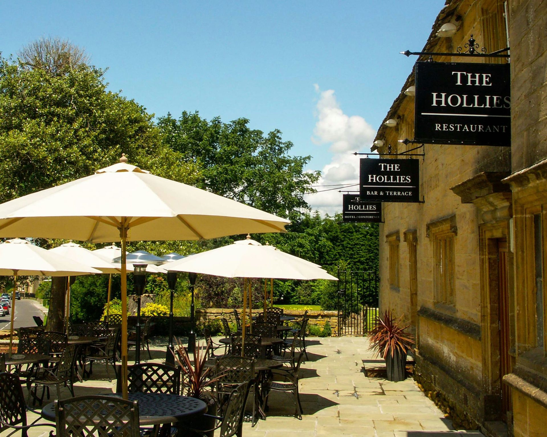 Outdoor dining terrace at The Hollies Hotel with cream umbrellas and traditional stone building.