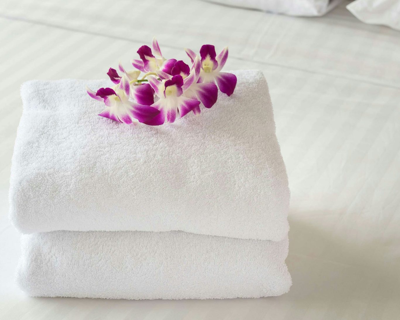 Stack of clean white towels topped with purple orchids on hotel bed.