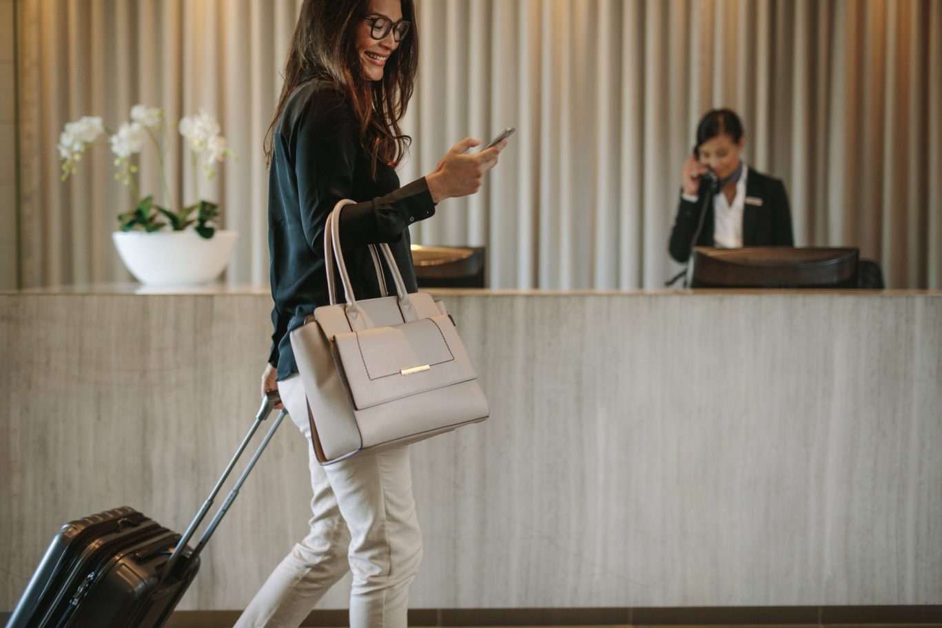 Smiling woman pulling her suitcase through a hotel lobby while checking her mobile phone.