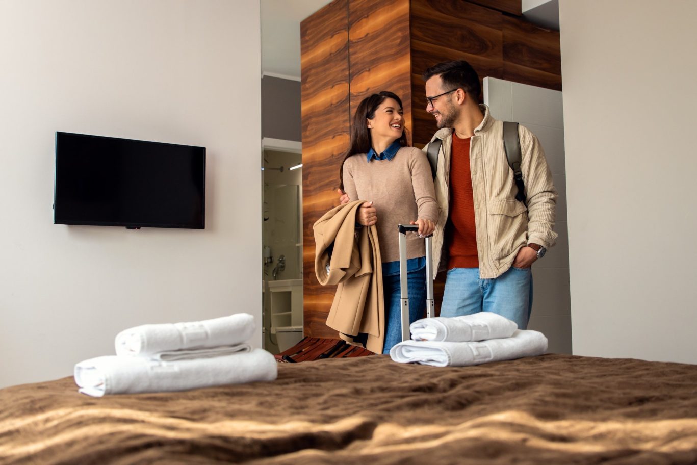 Smiling couple arriving at stylish hotel room with luggage and fresh towels on bed.