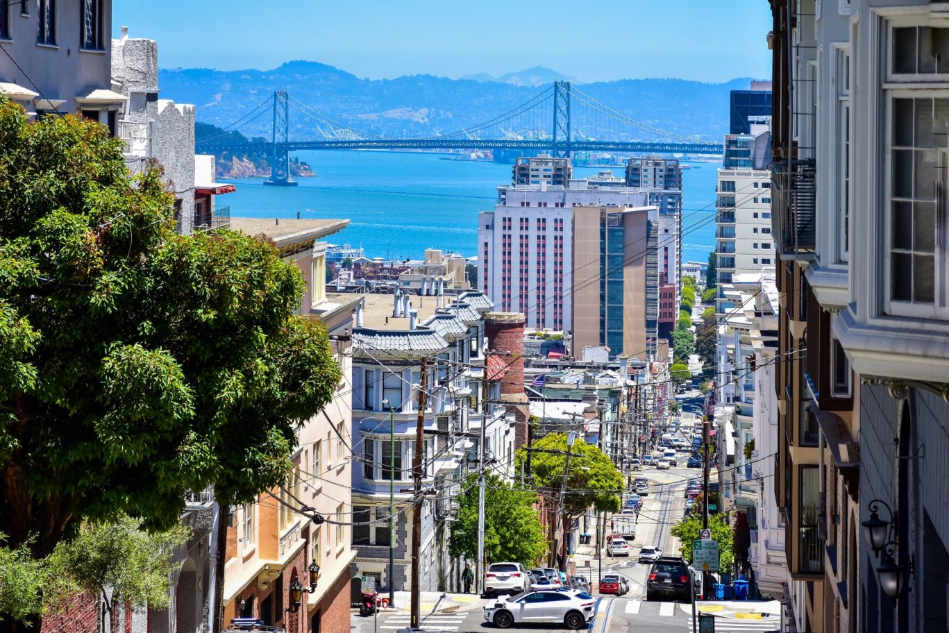 Steep San Francisco street descending toward the Bay Bridge with colorful buildings and ocean view.