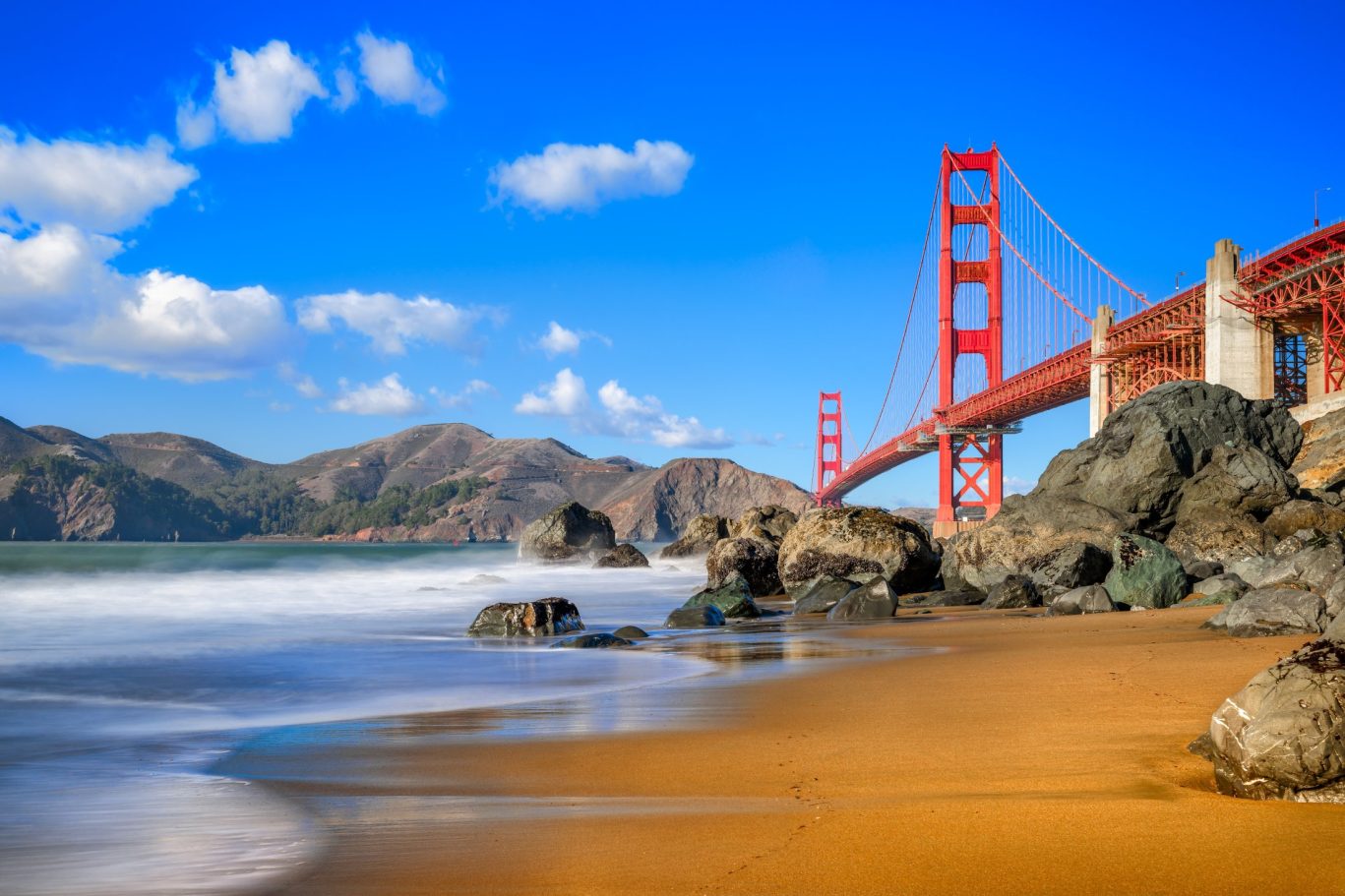 Golden Gate Bridge view from Baker Beach with rocky shoreline and blue sky.